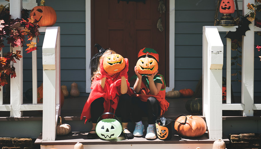 two young kids holding jack-o-lanterns by their heads sitting on their front porch