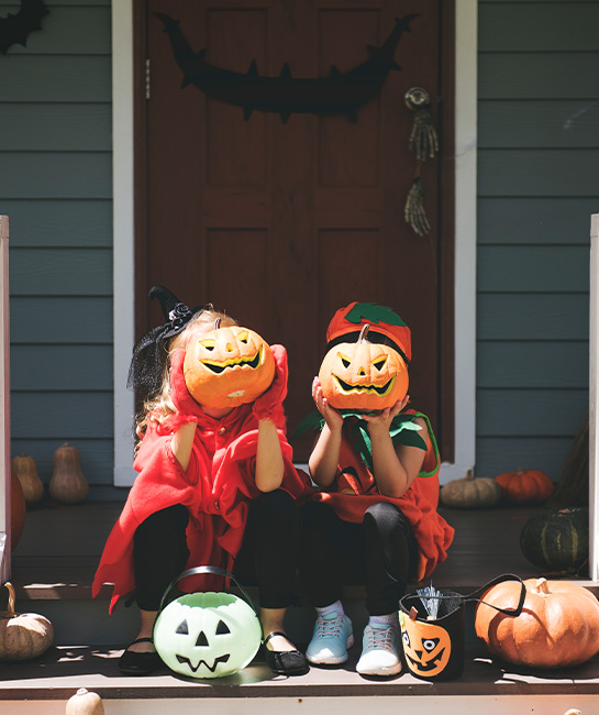 two young kids holding jack-o-lanterns by their heads sitting on their front porch