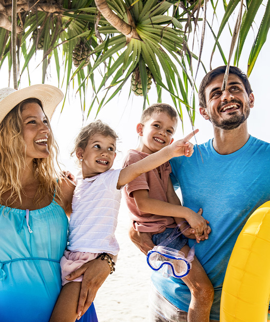 Family of four walking on the beach on vaction smiling holding kids and beach toys