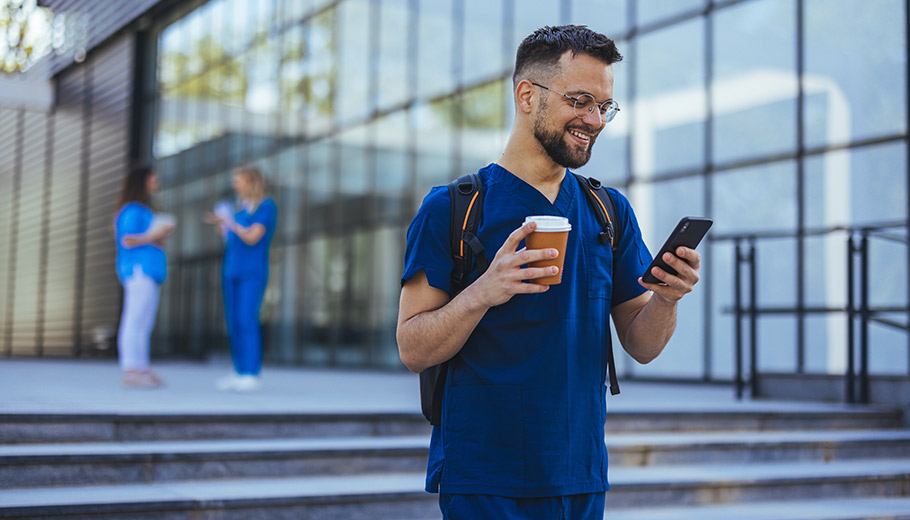 Male medical professional in blue scrubs walking outside smiling down at his phone