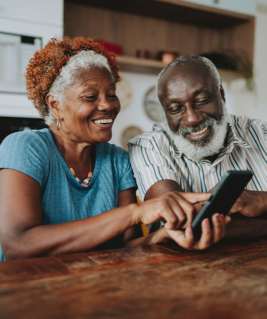 Older couple smiling and looking at a cell phone at their kitchen table
