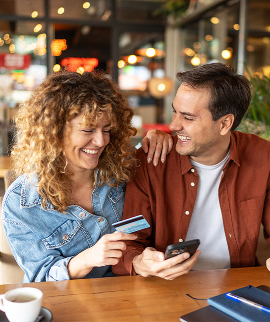 Couple at table looking at their cell phone smiling and holding a credit card