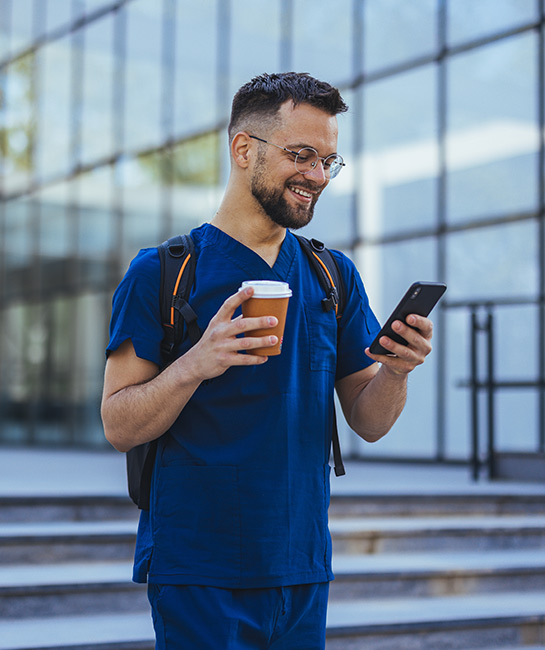 Male medical professional in blue scrubs walking outside smiling down at his phone